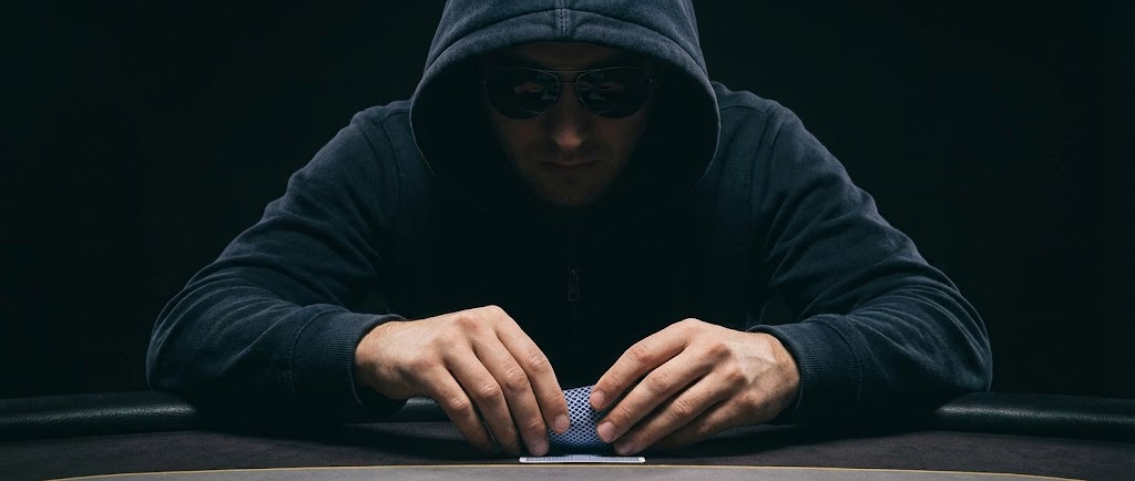 어두운 포커룸에서 검은색 후드와 선글라스를 착용한 플레이어가 테이블에 엎드려 자신의 홀 카드를 은밀하게 확인하는 손동작을 클로즈업한 와이드 페이지 배너 이미지.